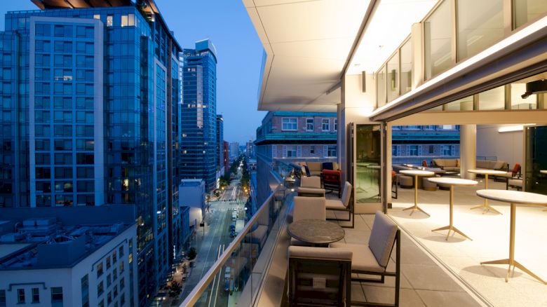 A modern balcony with furniture overlooks a street lined with tall buildings at twilight. The interior space features tables and tall stools.
