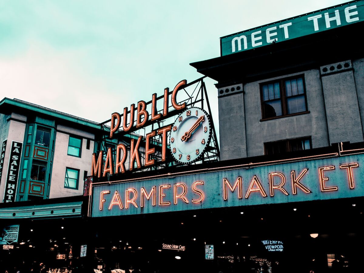 The image shows the iconic "Public Market Center" sign with a clock and a neon "Farmers Market" sign, indicating a popular market area.