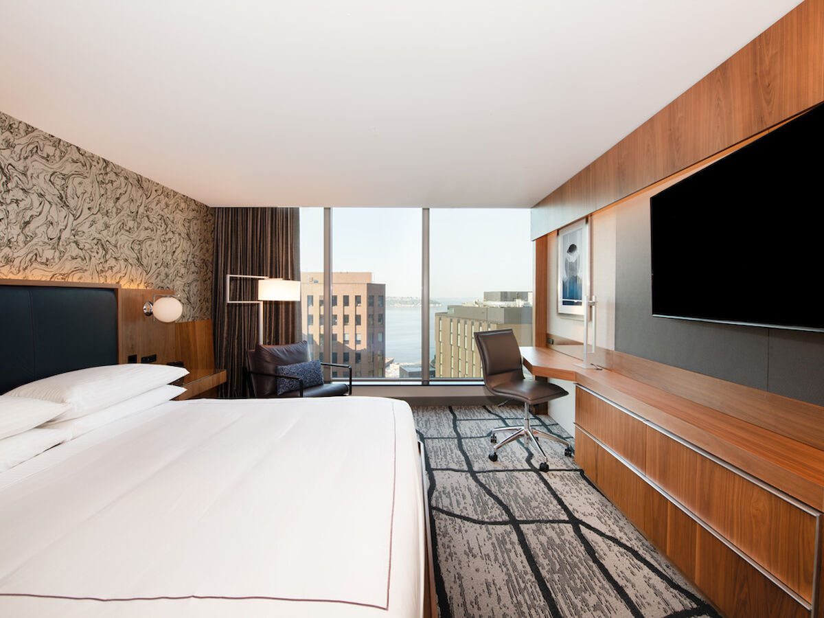 The image shows a modern hotel room with a large bed, a desk, a chair, a flat-screen TV, and a view of urban buildings through floor-to-ceiling windows.