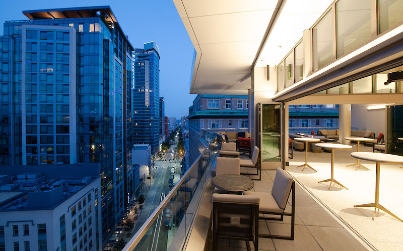 A modern balcony overlooking a city street with high-rise buildings, featuring outdoor seating and indoor areas with tables and chairs.