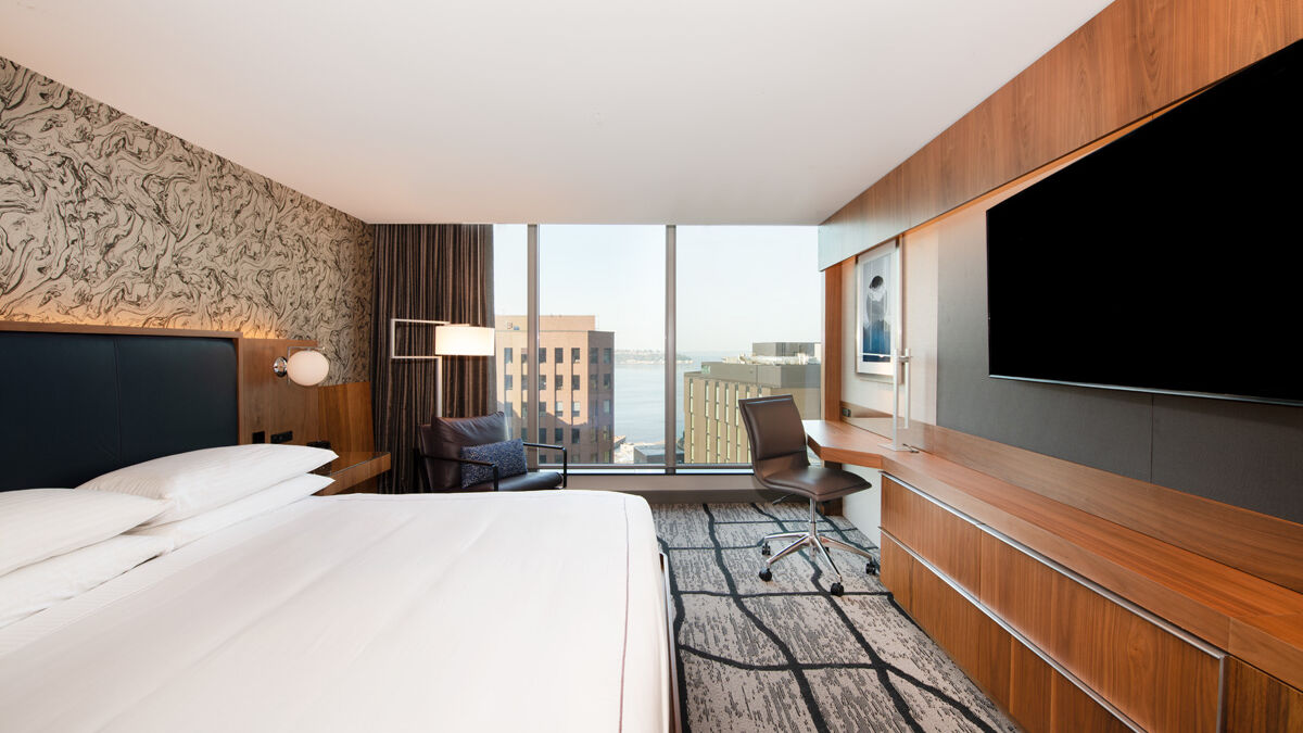 A modern hotel room features a large bed, wall-mounted TV, work desk, and a view of buildings and water through large windows.