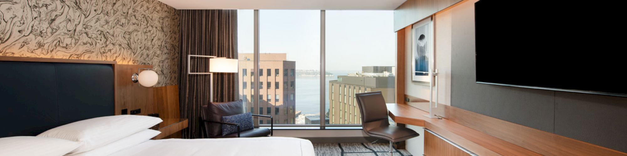 A modern hotel room features a large bed, wall-mounted TV, work desk, and a view of buildings and water through large windows.