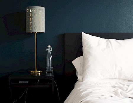 A neatly made bed with white pillows and a blanket, next to a black nightstand with a lamp and water bottle, against a dark blue wall.