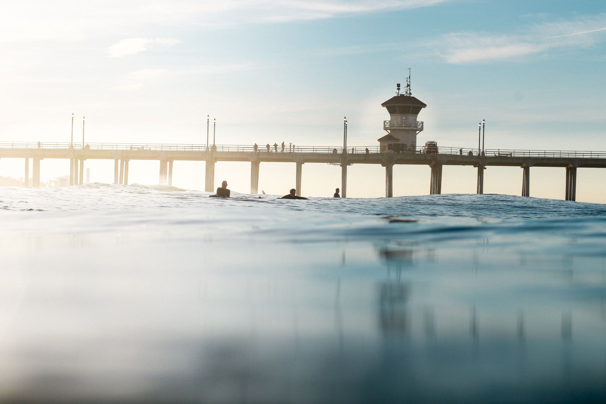 A serene coastal scene featuring a long pier, a watchtower, and a few people surfing or swimming in the gentle ocean waters.